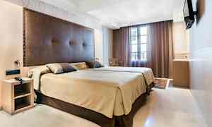 Hotellihuone, jossa suuri vuode, ruskea sängynpääty ja beige vuodevaatteet. Televisio on asennettu seinään, ja ikkunan peittää tummat verhot. (Tekoälyn luoma teksti)