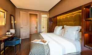 Hotellihuone, jossa on suuri sänky valkoisin vuodevaattein, kirjoituspöytä istuimineen ja ovi, joka johtaa kylpyhuoneeseen. (Tekoälyn luoma teksti)