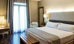 Hotellihuone, jossa on parivuode, kirjoituspöytä tuoleineen, nojatuoli ja suuret verhoilla varustetut ikkunat. (Tekoälyn luoma teksti)