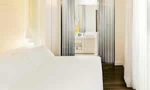 Hotellihuone, jossa on valkoinen sänky ja moderni kylpyhuone, näkyy pystysuorien mustien paneelien kautta. (Tekoälyn luoma teksti)