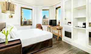 Hotellihuone, jossa on parivuode, tumma puulattia ja työpöytä televisiolla. Huoneessa on suuret ikkunat, joista avautuu kaunis näkymä kaupungille. (Tekoälyn luoma teksti)