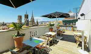 Kattoterassi, jossa on istuimia ja aurinkovarjoja, näkymä kaupungin vanhoille rakennuksille ja taustalla baarialue. (Tekoälyn luoma teksti)