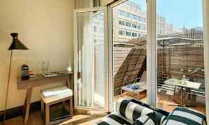 Huoneen sisustus, jossa kirjoituspöytä, tuoli ja avoin lasiovi, näkymä terassille, jossa kalusteita. (Tekoälyn luoma teksti)