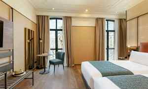 Moderni hotellihuone, jossa on kaksi sänkyä, pöytä ja tuoli suurten ikkunoiden edessä sekä puulattia. (Tekoälyn luoma teksti)