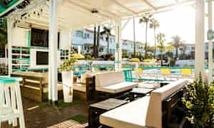 Ulko-oleskelualue valkoisen pergolan alla, jossa on huonekaluja ja kasveja, näkymä uima-altaalle ja ympäröiviin rakennuksiin. (Tekoälyn luoma teksti)