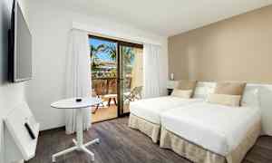 Moderni hotellihuone, jossa kaksi erillistä sänkyä, pieni pöytä ja seinälle asennettu televisio. Huoneessa on vaaleat värit ja ikkunasta pääsee parvekkeelle, jolta näkyy palmuja. (Tekoälyn luoma teksti)