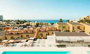 Kuva lomakohteesta, jossa on keltaisia rakennuksia, uima-allas, aurinkotuoleja ja merinäköala etäisyydellä. (Tekoälyn luoma teksti)