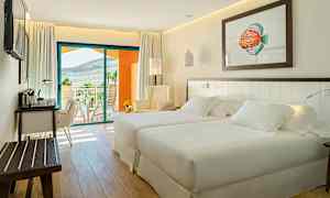 Valoisa ja moderni hotellihuone, jossa on kaksi sänkyä, oleskelualue ja kirjoituspöytä. Taustalla näkyy parveke maisemanäkymällä. (Tekoälyn luoma teksti)