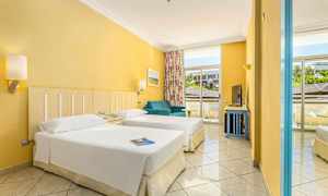 Hotellihuone, jossa on kaksi erillistä vuodetta, sininen sohva ja suuret ikkunat näkymällä ulos. (Tekoälyn luoma teksti)