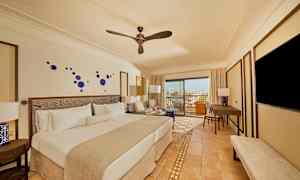 Hotellihuone, jossa on parivuode, istuinryhmä, työpöytä ja televisio. Huone on modernisti sisustettu sinisillä seinäkoristeilla, ja ikkunasta pääsee parvekkeelle, jolta on näköala. (Tekoälyn luoma teksti)