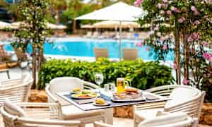 Pöytä, jolla on ruokaa ja juomaa, sijaitsee ulkoterassilla uima-altaan lähellä, ympärillä kukkivia kasveja ja aurinkovarjoja. (Tekoälyn luoma teksti)