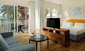 Kuva hotellihuoneesta, jossa on suuri sänky, sohva, pöytä ja televisio. Parveke, jossa on pöytä ja tuoleja, näkyy liukuovien kautta. (Tekoälyn luoma teksti)