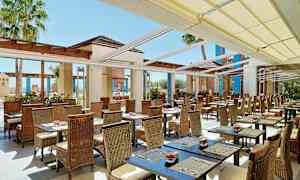 Ulkoterassi, jossa on paljon pöytiä ja tuoleja katoksen alla. Taustalla näkyy palmuja. (Tekoälyn luoma teksti)