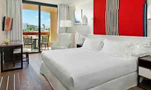 Hotellihuone, jossa on suuri sänky, nojatuoli, pöytä tuoleineen ja parveke, jolta on näkymä puutarhaan. (Tekoälyn luoma teksti)