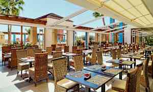 Ulkoterassi, jossa on rottinkituoleja ja pöytiä. Katosta peittää aurinkosuojus, ja taustalla näkyy palmuja. (Tekoälyn luoma teksti)