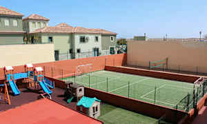 Kuvassa on ulkoleikkipaikka, jossa on liukumäki ja leikkikoteja sekä vihreä aidattu urheilukenttä jalkapalloa ja lentopalloa varten. Taustalla näkyy asuinrakennuksia. (Tekoälyn luoma teksti)