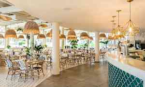 Kuva valoisasta ja modernista ravintolasta, jossa on valkoisia tuoleja ja pöytiä, luonnollisen tyylisiä kattovalaisimia ja baari pehmein linjoin. (Tekoälyn luoma teksti)