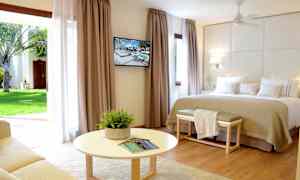 Valoisa hotellihuone, jossa on suuri sänky, seinälle kiinnitetty televisio ja istuinryhmä pyöreän pöydän edessä. Suuret verhot, jotka avautuvat näkymään puutarhaan. (Tekoälyn luoma teksti)