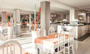 Ravintola, jossa on vaaleat kalusteet, pöydät pöytäliinoilla sekä koristeellisia elementtejä, kuten värikkäitä tankoasennelmia. (Tekoälyn luoma teksti)