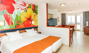 Hotellihuone, jossa sänky, värikäs kukkamaalaus seinällä sekä oleskelutila ja keittiö. (Tekoälyn luoma teksti)