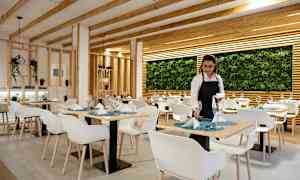 Ravintola, jossa on moderni sisustus, valkoiset tuolit, katetut pöydät ja seinä, jota koristavat kasvit. (Tekoälyn luoma teksti)