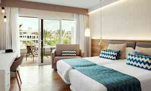 Hyvin valaistu hotellihuone, jossa on vuoteet, siniset koristeelliset tyynyt ja lasiovi, joka johtaa parvekkeelle näkymällä palmuihin. (Tekoälyn luoma teksti)