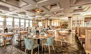 Tyylikäs ravintola-alue, jossa pyöreät pöydät ja kuviolliset tuolit, valaistuna kattovalaisimilla ja luonnonvalolla suurista ikkunoista. (Tekoälyn luoma teksti)