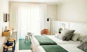 Hotellihuone, jossa on vaaleat seinät, kaksi sänkyä vihreillä peitteillä, pieni istuinryhmä tuoleilla ja pöydällä sekä seinälle kiinnitetty TV. (Tekoälyn luoma teksti)