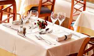 Katettu pöytä valkoisilla liinoilla, laseilla, aterimilla, lautasliinoilla ja pienellä kukkakoristeella. (Tekoälyn luoma teksti)