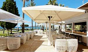 Ulkoterassi, jossa on pöytiä, tuoleja ja suuria aurinkovarjoja, näkymä puutarhaan ja merelle. (Tekoälyn luoma teksti)