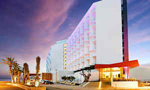 Moderni hotelli, jonka pääsisäänkäynnillä on valaistuja palmuja ja värikäs julkisivu. (Tekoälyn luoma teksti)