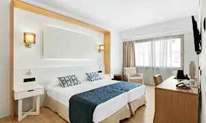 Hotellihuone, jossa kaksi sänkyä sinisillä päiväpeitteillä ja kuvioiduilla tyynyillä, työpöytä, tuoli sekä lepotuoli ikkunan vieressä. (Tekoälyn luoma teksti)