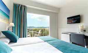 Hotellihuone, jossa on kaksi erillistä sänkyä, seinän kuvassa rantamaisema, suuret ikkunat, joista näkyy maisema, ja seinällä televisio. (Tekoälyn luoma teksti)