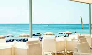 Näkymä rantakahvilasta, jossa on valkoisia tuoleja ja pöytiä, taustalla näkyy aurinkovarjoja ja meri. (Tekoälyn luoma teksti)