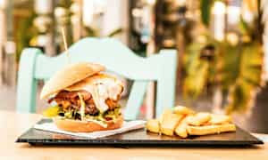 Hampurilainen ja lisukkeena ranskalaisia perunoita lautasella, asetettuna pöydälle taustalla tuoli. (Tekoälyn luoma teksti)