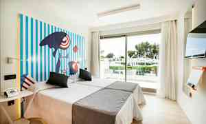 Valoisa hotellihuone, jossa on kaksi sänkyä, värikäs taide seinällä, lasiovi, josta avautuu näkymä vehreään puutarhaan, ja moderni televisio. (Tekoälyn luoma teksti)