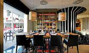 Modernin ravintolan sisustus, jossa on puupöytiä ja mustia tuoleja, ympäröitynä raidallisilla seinäkoristeilla ja suurilla ikkunoilla. (Tekoälyn luoma teksti)