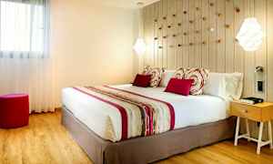 Makuuhuone, jossa on suuri sänky, koristetyynyjä ja värikoordinoitu päiväpeitto. Sängyn takana seinällä on koristeellisia yksityiskohtia, ja oikealla on yöpöytä. (Tekoälyn luoma teksti)