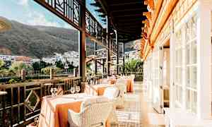 Ravintola, jossa on pöytiä ja tuoleja terassilla, näkymä rakennuksiin ja vuorille. (Tekoälyn luoma teksti)