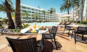 Ulkotila uima-altaan äärellä, jossa on aurinkotuoleja ja varjoja. Pöytä, jossa on ruokaa ja juomaa, on etualalla. (Tekoälyn luoma teksti)