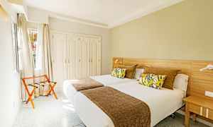 Hotellihuone, jossa on kaksi yhden hengen vuodetta, koristeelliset tyynyt ja ruskea päiväpeite, vaatekaappi ja tuoli ikkunan vieressä. (Tekoälyn luoma teksti)