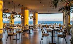 Kuva ravintolasta, jossa on rottinkituoleja, pöydät ovat katettuina ja merimaisema näkyy suurten lasi-ikkunoiden läpi. (Tekoälyn luoma teksti)
