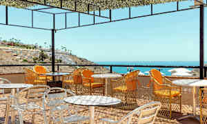 Ulkoterassi, jossa pöytiä ja tuoleja, näkymä merelle ja rinteeseen rakennuksineen. (Tekoälyn luoma teksti)