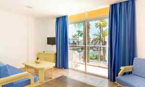 Valoisa hotellihuone, jossa on siniraidallinen sohva, pöytä, jonka päällä on samppanjapullo ja laseja, pieni televisio tason päällä sekä suuret ikkunat sinisin verhoin, joita kautta pääsee parvekkeelle näkymällä palmuja. (Tekoälyn luoma teksti)