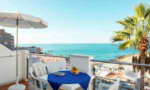 Näkymä parvekkeelta, jossa on sinisellä liinalla peitetty pöytä ja ympärillä tuoleja. Pöydällä on hedelmäkori, ja taustalla näkyy ranta, palmuja ja meri. (Tekoälyn luoma teksti)