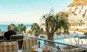 Henkilö istuu terassilla, josta on näkymä merelle, puille ja rakennuksille, mukanaan kahvikuppi ja aamiainen. (Tekoälyn luoma teksti)