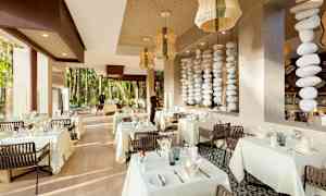 Tyylikäs ravintola, jossa on valkoiset pöytäliinat, koristellut pöydät ja suuret ikkunat viheralueelle. (Tekoälyn luoma teksti)