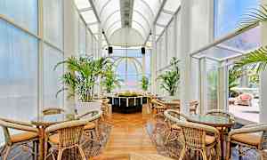 Korikalusteet ja pöydät korkeassa kaarisella katolla, joita ympäröivät suuret lasiosiot ja vihreät kasvit. (Tekoälyn luoma teksti)