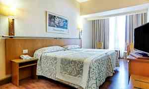 Hotellihuone, jossa on parivuode kuviollisilla päiväpeitteillä, seinävalaisimia ja televisio kirjoituspöydällä. Istuimia ja verhoilla varustettu ikkuna ovat myös mukana. (Tekoälyn luoma teksti)