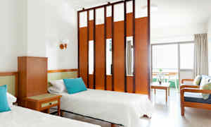 Hotellihuone, jossa on kaksi yhden hengen vuodetta, puinen pöytä ja istuinryhmä, joita erottaa huoneiden jakaja. (Tekoälyn luoma teksti)